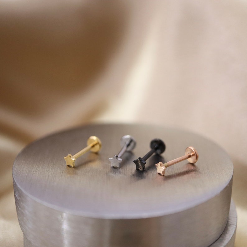 Puede incluir: Cuatro anillos de nariz en forma de estrella en diferentes colores: oro, plata, negro y oro rosa. Los anillos se muestran en una superficie plateada.