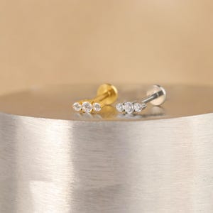 K&ouml;nnte beinhalten: Zwei kleine, minimalistische Ohrringe mit jeweils drei klaren Zirkonia-Steinen. Die Ohrringe liegen auf einer silbernen und goldenen Metalloberfl&auml;che.