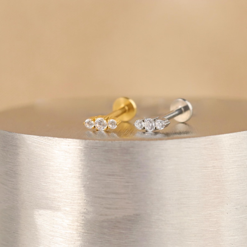 K&ouml;nnte beinhalten: Zwei kleine, minimalistische Ohrringe mit jeweils drei klaren Zirkonia-Steinen. Die Ohrringe liegen auf einer silbernen und goldenen Metalloberfl&auml;che.