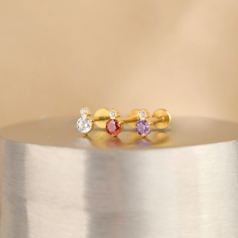 Puede incluir: Tres piezas de joyer&iacute;a corporal de color dorado con una peque&ntilde;a piedra transparente en la parte superior y una piedra de color m&aacute;s grande en la parte inferior. Las piedras son rojas, moradas y transparentes.