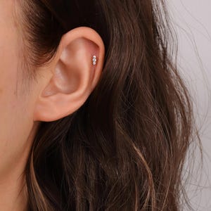K&ouml;nnte beinhalten: Nahaufnahme eines Ohrs mit einem kleinen, silbernen Ohrstecker mit drei winzigen, klaren Edelsteinen.
