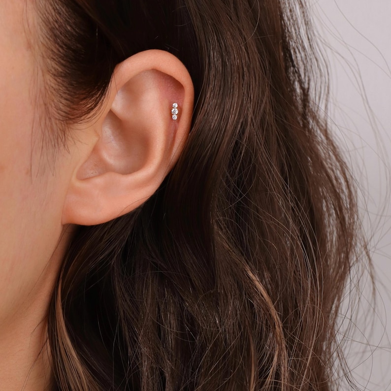 K&ouml;nnte beinhalten: Nahaufnahme eines Ohrs mit einem kleinen, silbernen Ohrstecker mit drei winzigen, klaren Edelsteinen.