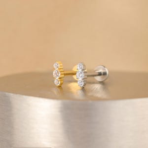 K&ouml;nnte beinhalten: Zwei kleine Ohrringe mit einer Reihe von drei klaren Zirkonia-Steinen. Der Ohrring links ist golden und der Ohrring rechts ist silber.