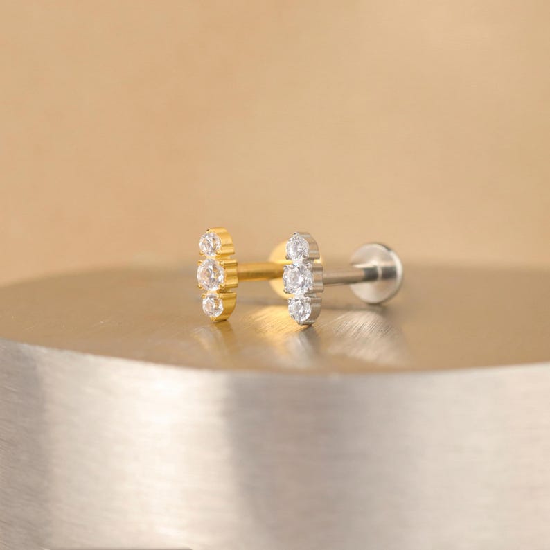 K&ouml;nnte beinhalten: Zwei kleine Ohrringe mit einer Reihe von drei klaren Zirkonia-Steinen. Der Ohrring links ist golden und der Ohrring rechts ist silber.