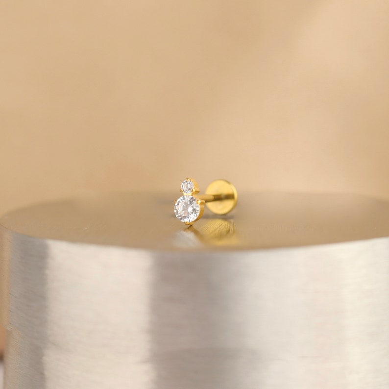 Puede incluir: Un pendiente de bot&oacute;n dorado con dos piedras de circonita c&uacute;bica transparentes. La piedra m&aacute;s grande es redonda y la m&aacute;s peque&ntilde;a est&aacute; colocada encima.