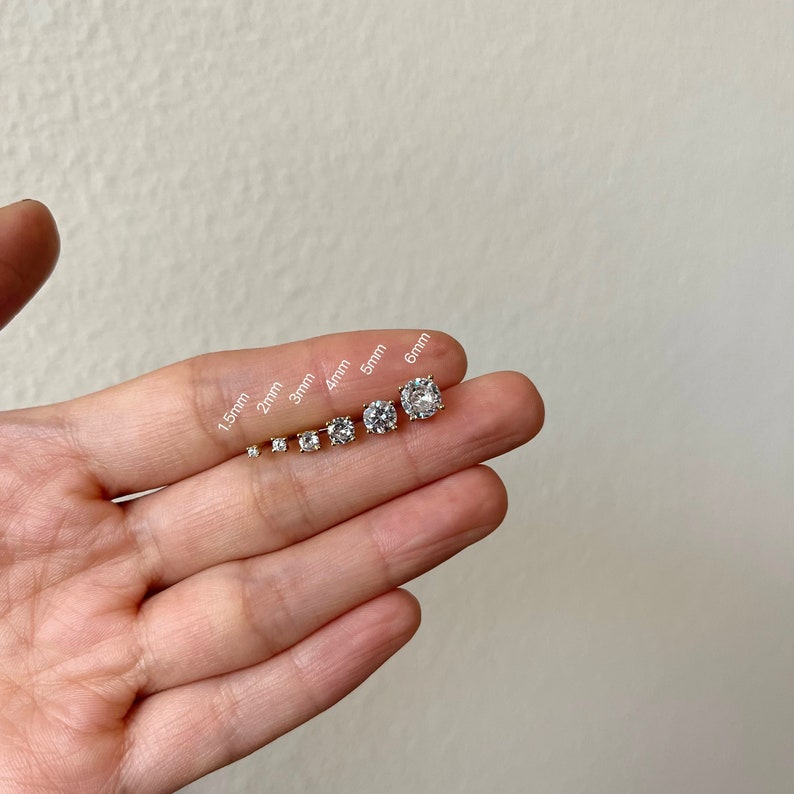 Pu&ograve; includere: Un set di sette orecchini a perno in zircone cubico trasparente di diverse dimensioni, che vanno da 1,5 mm a 6 mm, mostrati su una mano.
