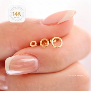 Puede incluir: Tres pendientes de botón dorados con forma de círculo abierto en diferentes tamaños. Los pendientes se sostienen entre los dedos, mostrando su delicado diseño. La imagen incluye un logotipo que dice "CEENIN JEWELRY 14K GOLD".