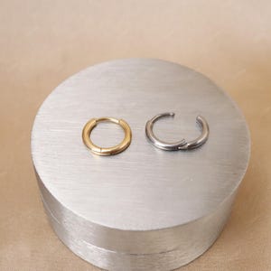 May include: Two small hoop earrings, one gold and one silver, are displayed on a silver, circular surface. The gold earring is closed, while the silver earring is open, showing the clasp. The earrings are simple and modern.