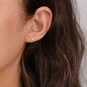 K&ouml;nnte beinhalten: Nahaufnahme eines Ohrs mit einem kleinen, zarten Ohrring mit drei Steinen. Der Ohrring ist aus Gold und hat drei winzige, klare Edelsteine.