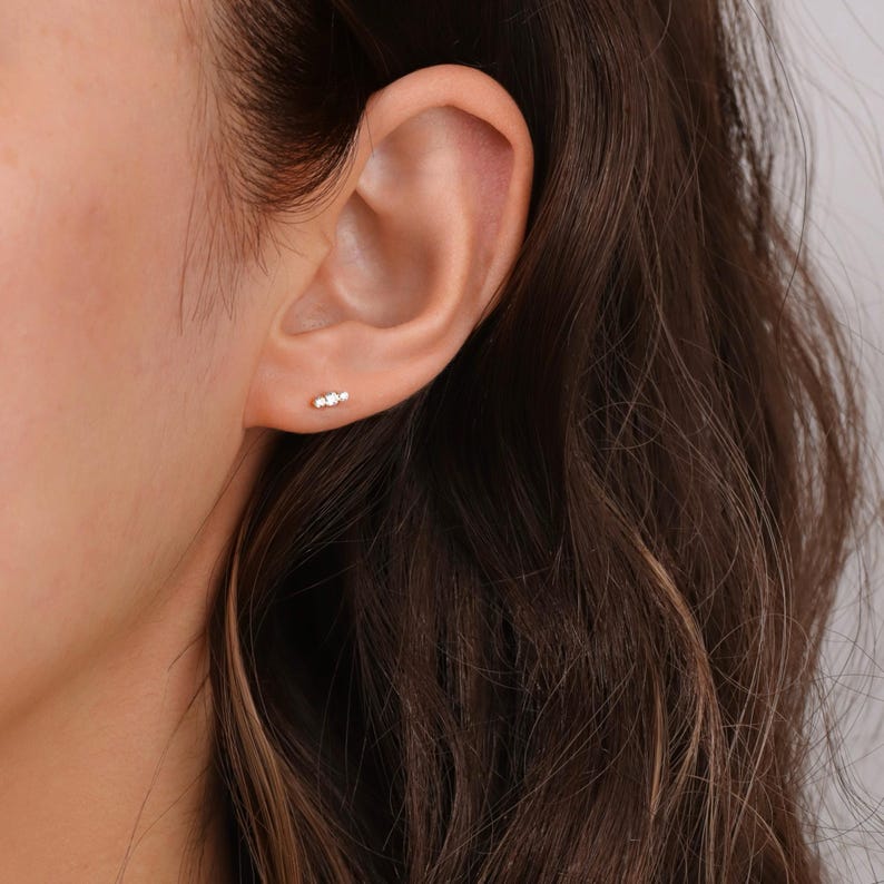 K&ouml;nnte beinhalten: Nahaufnahme eines Ohrs mit einem kleinen, zarten Ohrring mit drei Steinen. Der Ohrring ist aus Gold und hat drei winzige, klare Edelsteine.