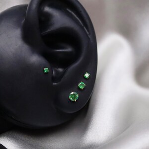 May include: Four green gemstone earrings on a black ear model. The earrings are different sizes and are set in gold.