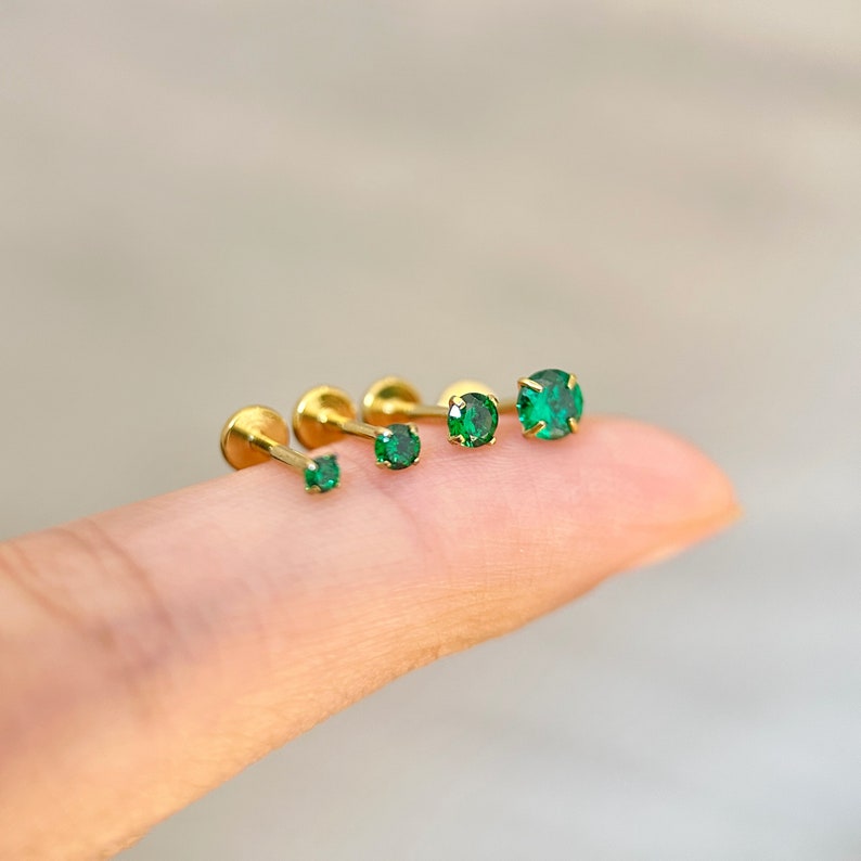 May include: Four gold body jewelry pieces with green gem stones. The pieces are lined up in order of size, with the smallest on the left and the largest on the right.