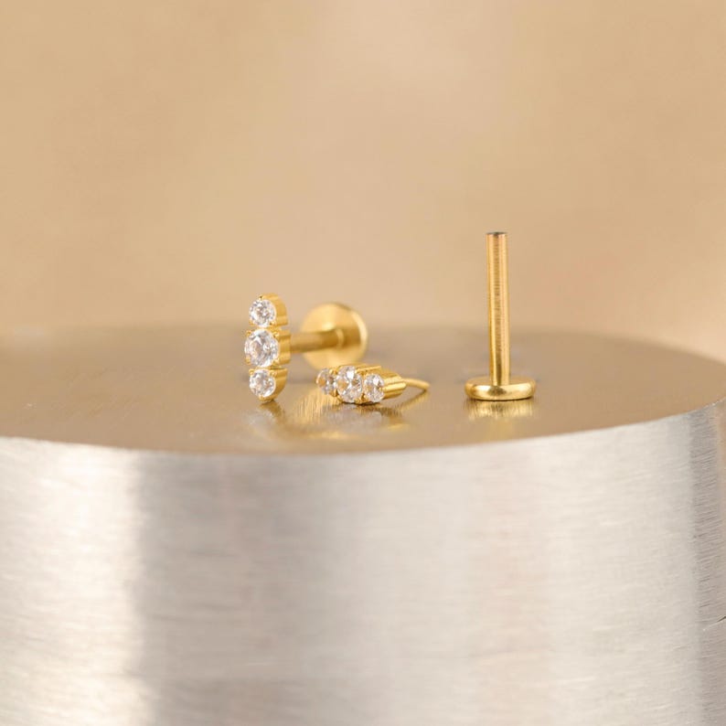 K&ouml;nnte beinhalten: Goldener K&ouml;rperschmuck mit einer Reihe von klaren Zirkonia-Steinen. Der Schmuck wird auf einer silbernen Oberfl&auml;che gezeigt.