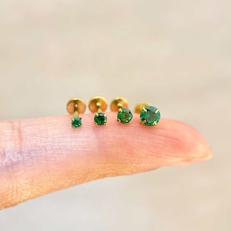 May include: Four gold body jewelry pieces with green gemstones. The pieces are arranged in order of size, with the smallest on the left and the largest on the right.