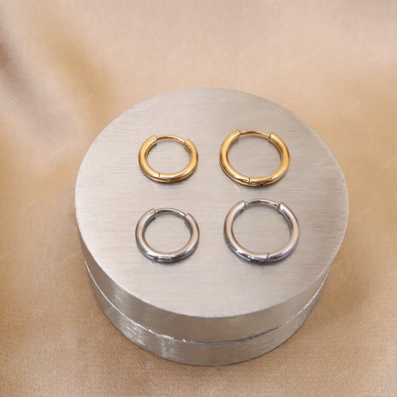 May include: Four hoop earrings, two gold-toned and two silver-toned, are displayed on a silver platform. The earrings are of varying sizes, with the gold hoops being larger than the silver ones. The background is a neutral beige.