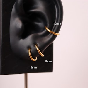 May include: Gold-colored hoop earrings, 8mm and 10mm in diameter, displayed on a black ear model. The earrings are simple and elegant, suitable for everyday wear. The image is set against a soft pink background.