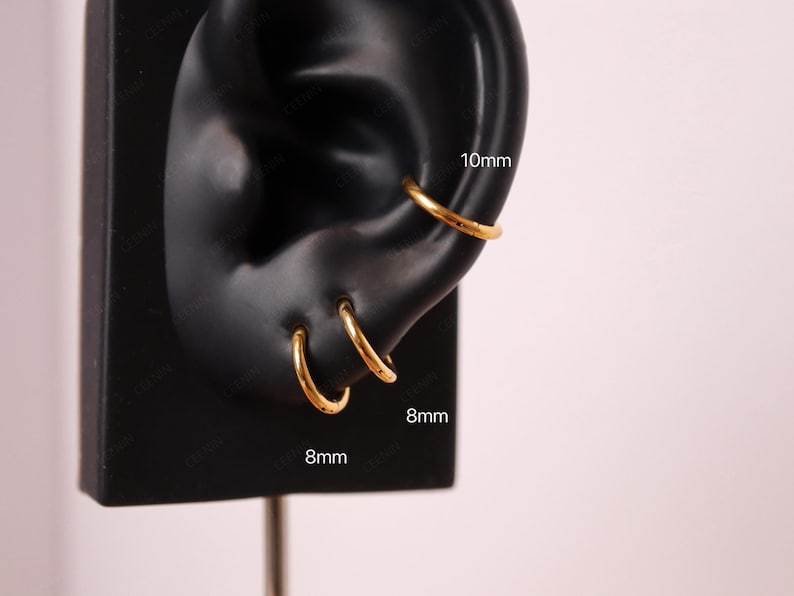 May include: Gold-colored hoop earrings, 8mm and 10mm in diameter, displayed on a black ear model. The earrings are simple and elegant, suitable for everyday wear. The image is set against a soft pink background.