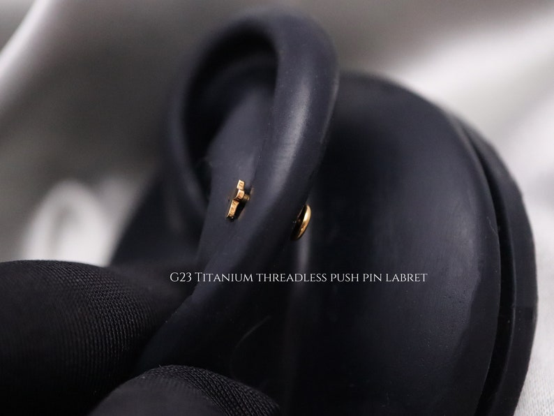 Puede incluir: Primer plano de un piercing de labret de titanio dorado sin rosca. El labret est&aacute; en un expositor negro.