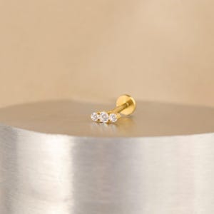 K&ouml;nnte beinhalten: Goldener K&ouml;rperschmuck mit einer Reihe von drei klaren Zirkonia-Steinen.