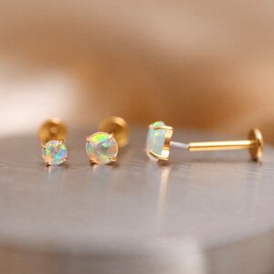 K&ouml;nnte beinhalten: Drei goldene Opal-Ohrringe, zwei sind Ohrstecker und einer ist ein Barbell-Ohrring. Die Opal-Steine sind irisierend und haben eine wei&szlig;e Basis.