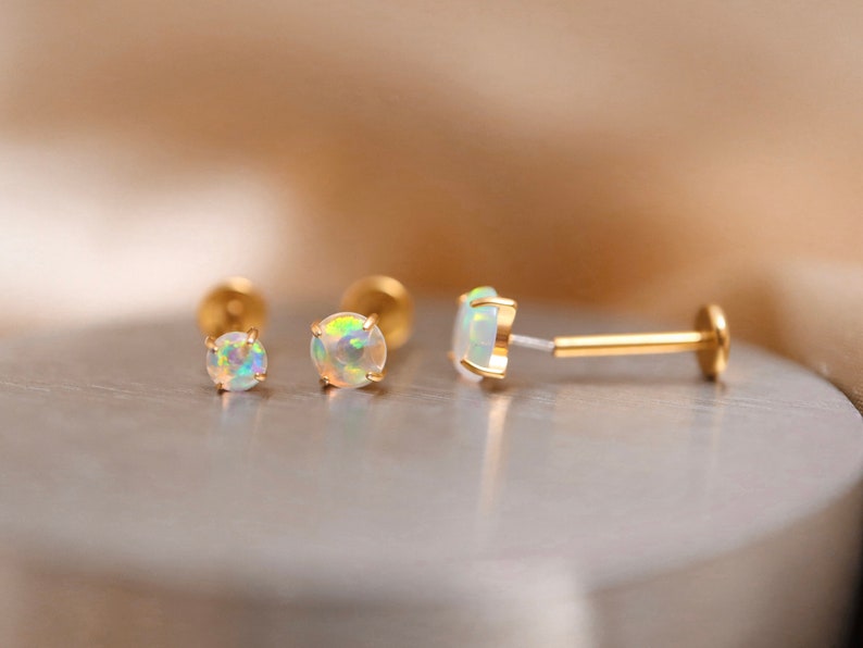 K&ouml;nnte beinhalten: Drei goldene Opal-Ohrringe, zwei sind Ohrstecker und einer ist ein Barbell-Ohrring. Die Opal-Steine sind irisierend und haben eine wei&szlig;e Basis.