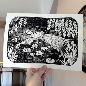 May include: A black and white linocut print on paper featuring a figure in a flowing dress floating in water. The scene includes water lilies, lily pads, tall grasses, and overhanging willow branches, all rendered in high-contrast ink lines.