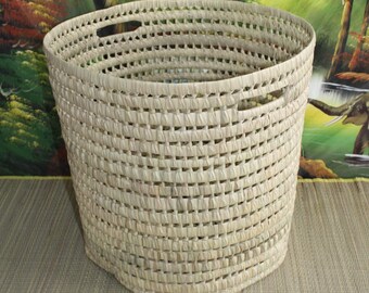 May include: A large, round, woven basket made of natural materials. The basket has a light brown color and is made with a tight weave. The basket has two handles on the side.