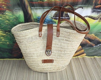 Puede incluir: Un gran bolso tote de paja tejido natural con asas de cuero marrón y cierre de hebilla. El bolso tiene una etiqueta que dice "Bhella".