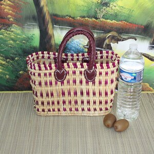 May include: A woven basket with a purple and natural straw pattern and brown leather handles. The basket is sitting on a woven mat with a bottle of water and two kiwi fruits in front of it.