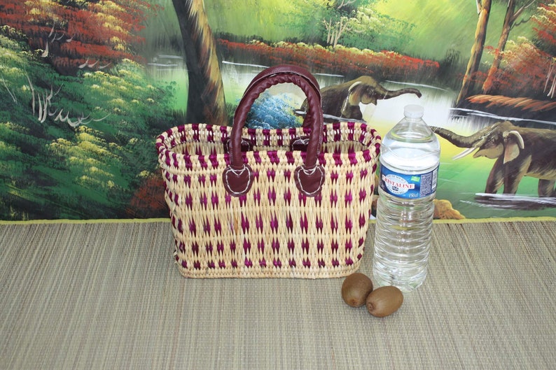 May include: A woven basket with a purple and natural straw pattern and brown leather handles. The basket is sitting on a woven mat with a bottle of water and two kiwi fruits in front of it.