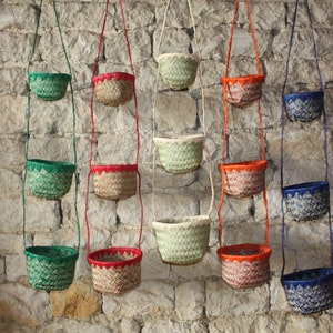 Può includere: Una collezione di piccoli cestini sospesi, tessuti, in vari colori, tra cui verde, rosso, arancione e blu. Ogni cestino è sospeso da un cordino abbinato contro un muro di pietra strutturato. I cestini presentano un motivo tessuto decorativo.
