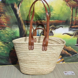 Puede incluir: Una cesta grande tejida de forma natural con dos asas largas de cuero marrón. La cesta está hecha de paja y tiene un color beige claro. Las asas están cosidas con hilo blanco y tienen un diseño decorativo.