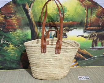 May include: A large, natural woven basket with two long brown leather handles. The basket is made of straw and has a light beige color. The handles are stitched with white thread and have a decorative design.