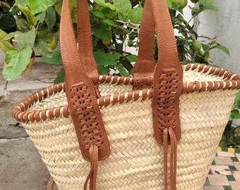 Puede incluir: Un bolso tote de paja tejido con asas de cuero marrón y detalles de flecos. El bolso tiene una base ancha y redondeada y una parte superior ligeramente curvada.