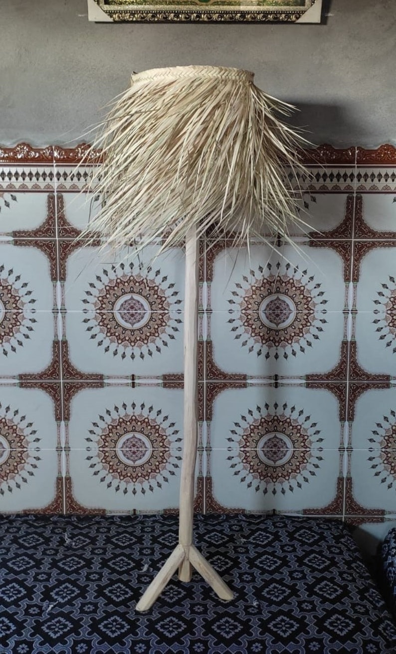 Puede incluir: Una pantalla de l&aacute;mpara alta tejida de forma natural con una franja de material similar a la paja. La pantalla de la l&aacute;mpara est&aacute; montada en un soporte de tr&iacute;pode de madera.