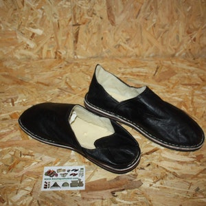 Op de afbeelding: Twee paar zwarte leren slippers met witte stiksels. De slippers hebben een gesloten neus en een platte zool.