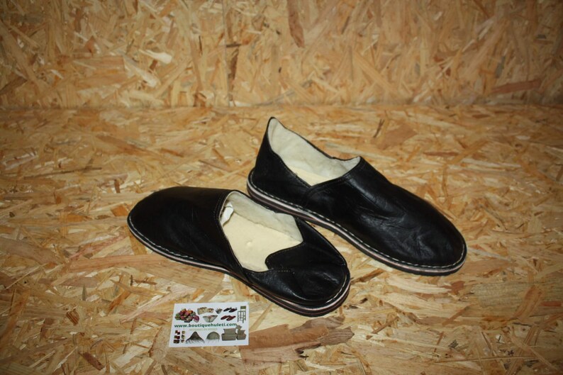 Op de afbeelding: Twee paar zwarte leren slippers met witte stiksels. De slippers hebben een gesloten neus en een platte zool.