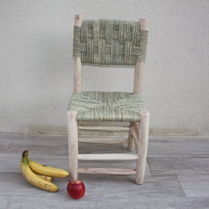 Pode incluir: Uma cadeira de madeira com assento e encosto de vime. A cadeira &eacute; de cor castanho claro e tem um acabamento natural. H&aacute; tr&ecirc;s bananas e uma ma&ccedil;&atilde; vermelha no ch&atilde;o &agrave; frente da cadeira.