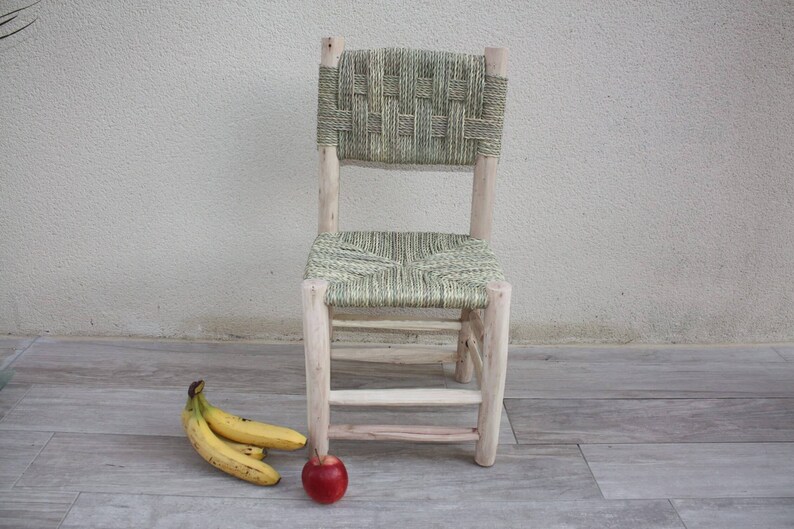Pode incluir: Uma cadeira de madeira com assento e encosto de vime. A cadeira &eacute; de cor castanho claro e tem um acabamento natural. H&aacute; tr&ecirc;s bananas e uma ma&ccedil;&atilde; vermelha no ch&atilde;o &agrave; frente da cadeira.