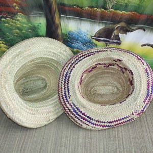 Puede incluir: Dos sombreros de paja tejidos, uno con un dise&ntilde;o liso y el otro con rayas moradas y blancas. Los sombreros est&aacute;n sobre una superficie marr&oacute;n.