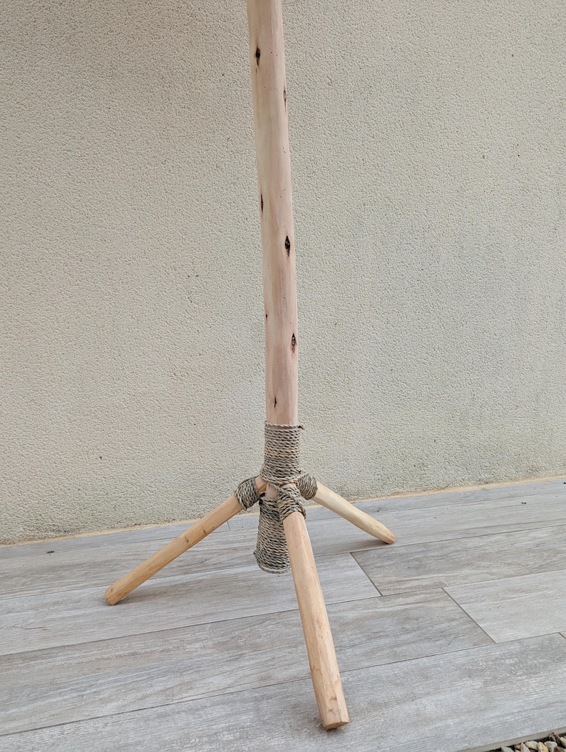 Puede incluir: Un soporte de tr&iacute;pode de madera con un acabado natural. El soporte tiene tres patas que est&aacute;n conectadas a un poste central con una envoltura de cuerda.