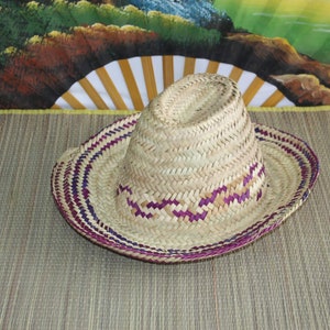 Puede incluir: Un sombrero de paja tejido con ribete morado y blanco. El sombrero est&aacute; sobre una estera tejida.