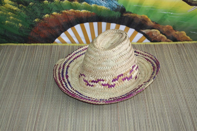 Puede incluir: Un sombrero de paja tejido con ribete morado y blanco. El sombrero est&aacute; sobre una estera tejida.