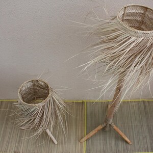 Puede incluir: Dos pantallas de l&aacute;mparas tejidas naturales con una textura similar a la paja. La pantalla m&aacute;s grande est&aacute; sobre un soporte de madera con tres patas. La pantalla m&aacute;s peque&ntilde;a est&aacute; sobre un soporte de madera con una pata.