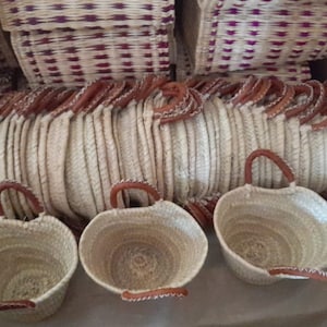 Pode incluir: Uma pilha de cestas de palha tecidas com alças de couro. As cestas são de cor castanho claro e têm um aspeto natural e rústico.