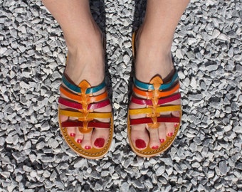 May include: A pair of colourful leather sandals with straps in shades of red, orange, yellow, blue, and green. The sandals have a simple design and are perfect for warm weather.