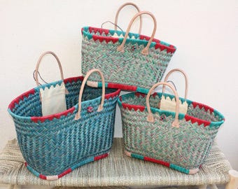 May include: Three woven tote bags in shades of teal and green, with red and teal trim. The bags have tan leather handles and a white fabric lining. The bags are arranged on a woven bench.