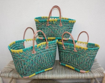 May include: A stack of four woven tote bags. The bags are green with yellow and brown accents. They have brown handles and are sitting on a woven bench. The bags are empty.