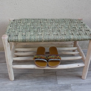 Op de afbeelding: Een houten bank met een gevlochten zitting en een plank eronder. Twee paar sandalen met kleurrijke kwastjes staan op de plank.