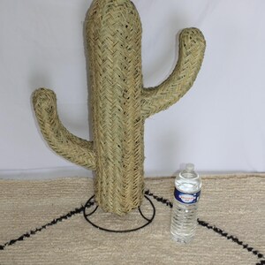 Puede incluir: Una gran escultura de cactus beige tejida con un soporte de metal negro. El cactus tiene dos brazos y est&aacute; de pie sobre una alfombra blanca con ribete negro.